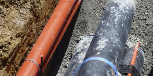 Open sleuf met een oranje rioleringsbuis en grote grijze leiding met blauwe banden op een werf van Amery bvba